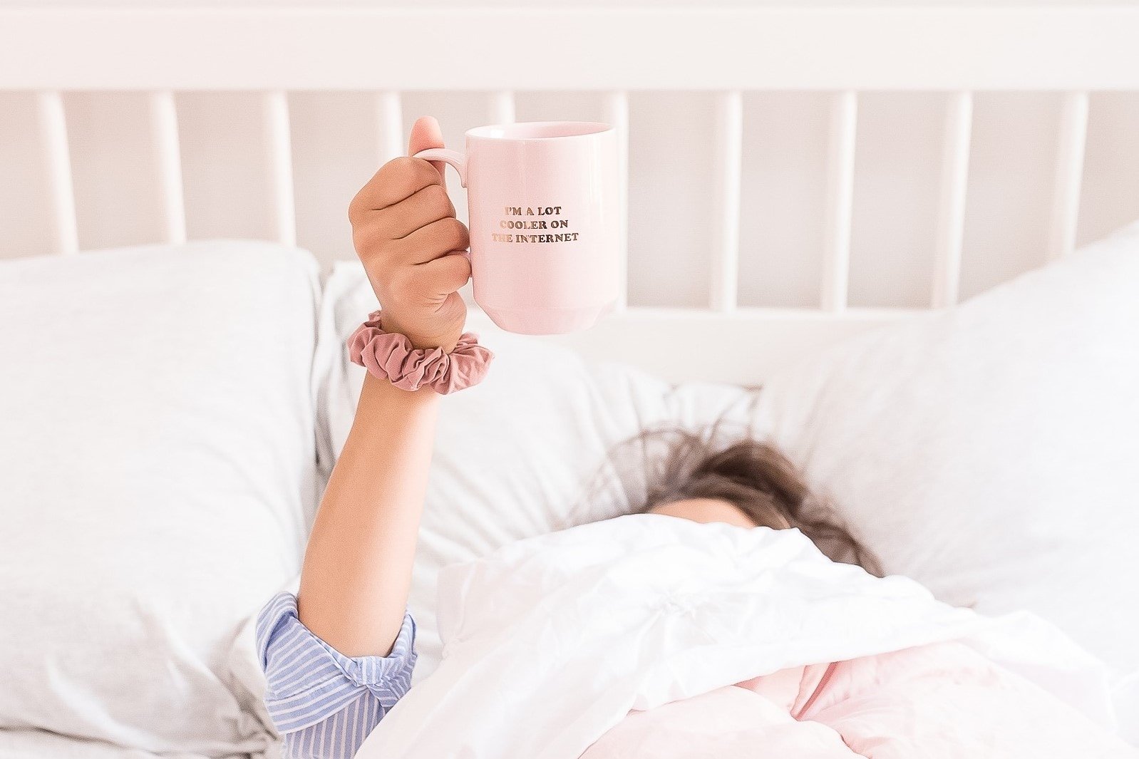 Woman in bed, covered by the duvet, holding up a mug which reads 'I am a lot cooler on the internet'