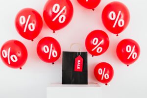 Small black bag with red 'sale' tag, surrounded by red balloons with % symbols on them